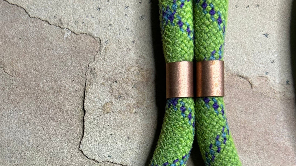 A close-up of a green braided rope with a blue and purple zigzag pattern, featuring two copper-colored metal spacers, laid on a textured stone surface.