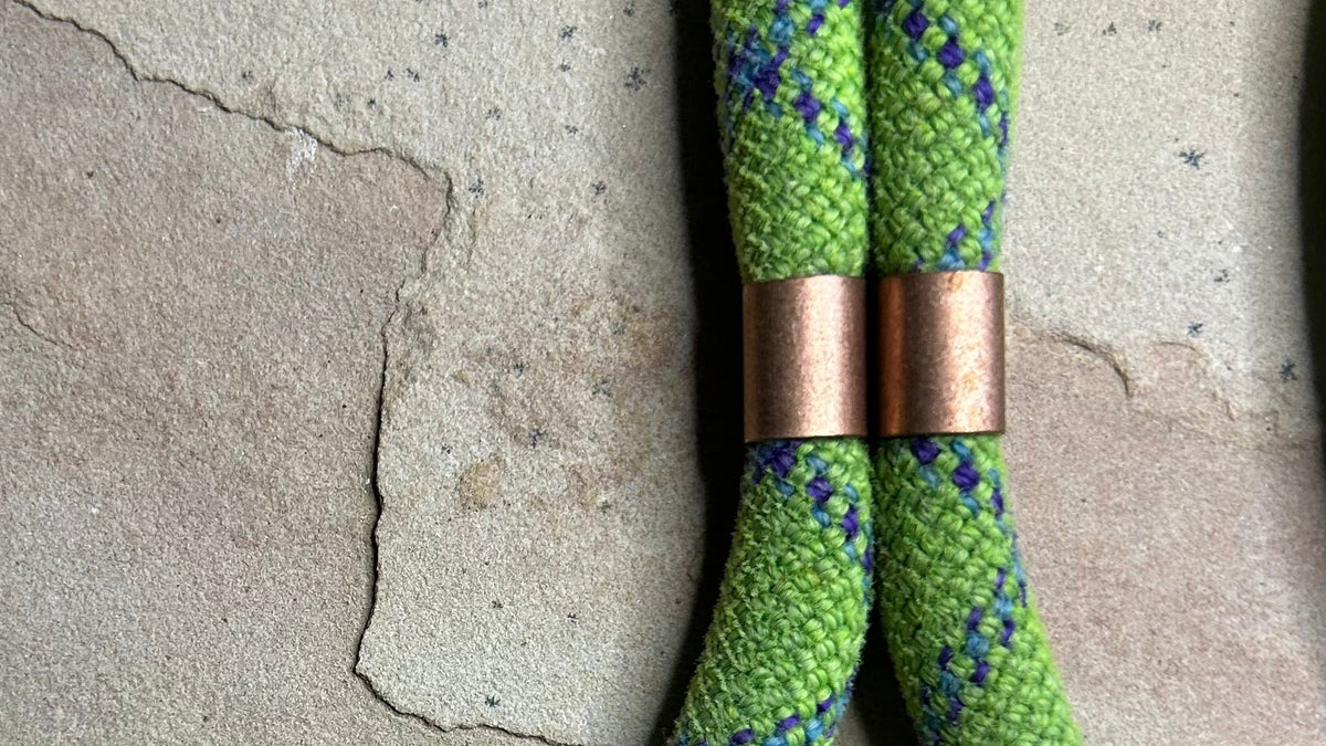 A close-up of a green braided rope with a blue and purple zigzag pattern, featuring two copper-colored metal spacers, laid on a textured stone surface.